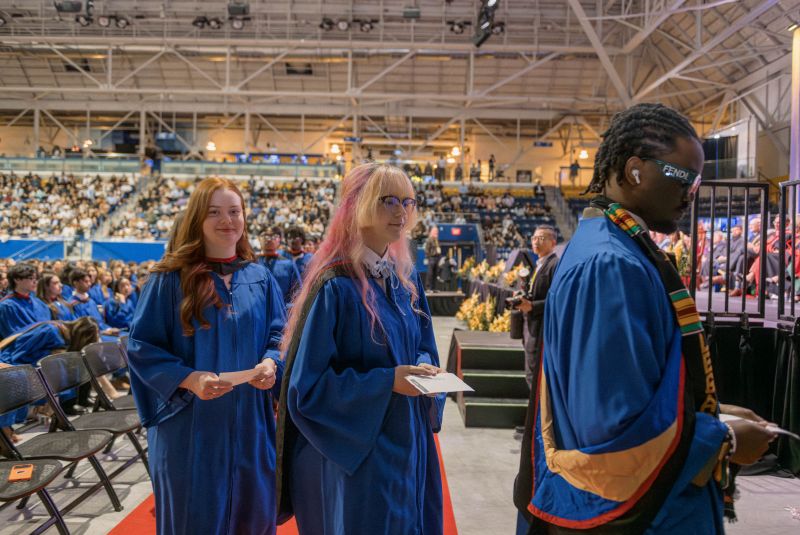 three grads in glue gowns are lined up to cross the stage and are holding their name reader cards