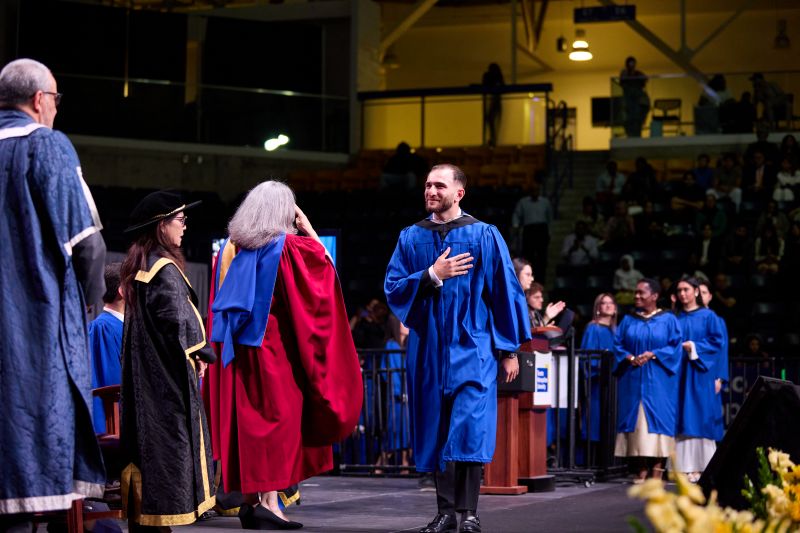 A grad in a blue gown places their hand over their heart as they cross the stage
