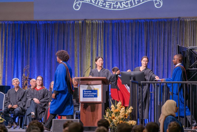 A grad in a blue gowns hands their name reader card to a staff member on stage in a black gown