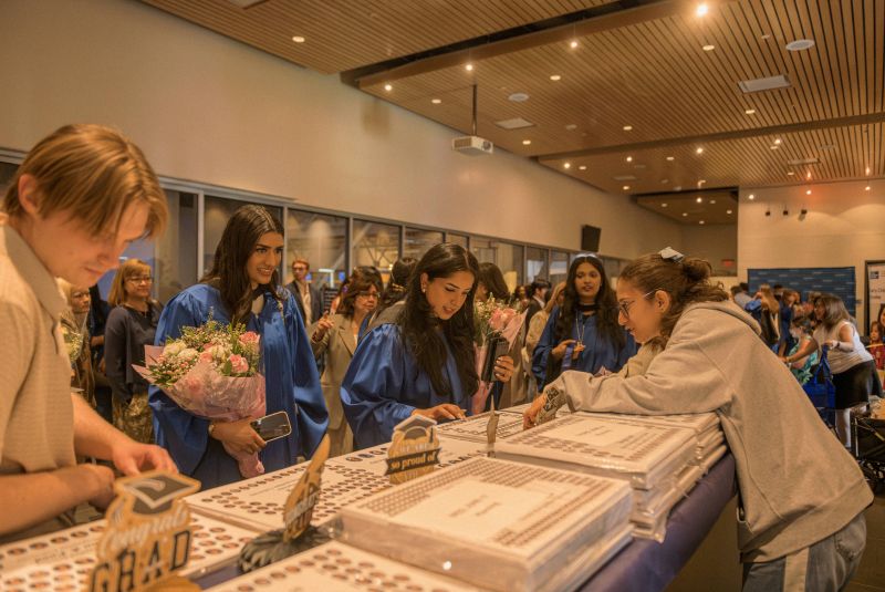 Grads in blue gowns are lined up in front of a table where class composites are being distributed
