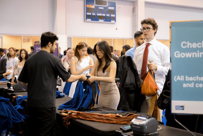 A grad is handing a staff member a blue gown at a gown return station in the Varsity Court