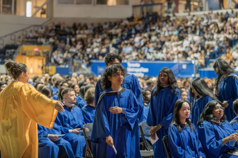 Grads in blue gowns are standing in a single file line on the ramp leading to the stage
