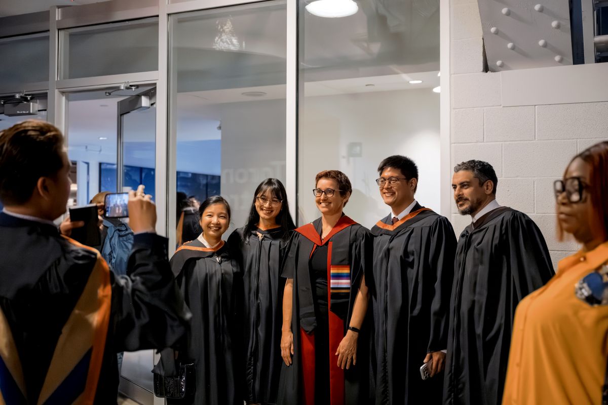 A group of graduands in black masters gowns with a faculty member in a black gown and rainbow LGBTQIA2S+ stole