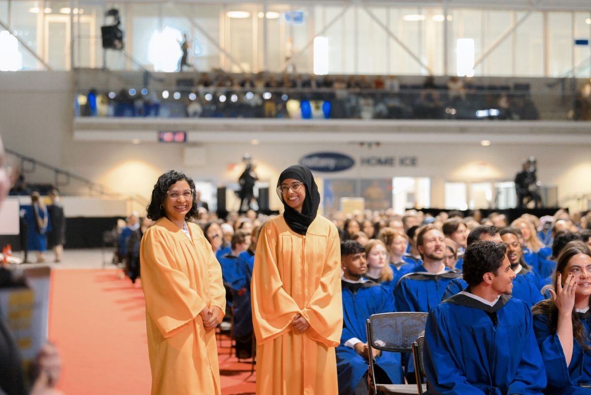 Two smiling convocation staff members in gold gowns are standing in the Convocation arena surrounded by seated graduates wearing blue Bachelor's gown. 
