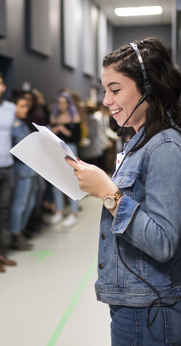 A student wearing a headset holds a call sheet at a modeling audition with the models standing in a line.