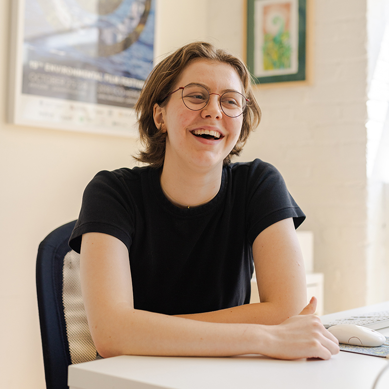 A person sitting at a desk in a bright office, smiling while working at a computer.