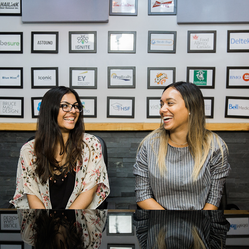 Two people seated at a table in a professional setting, smiling and talking, with company logos displayed on the wall behind them.