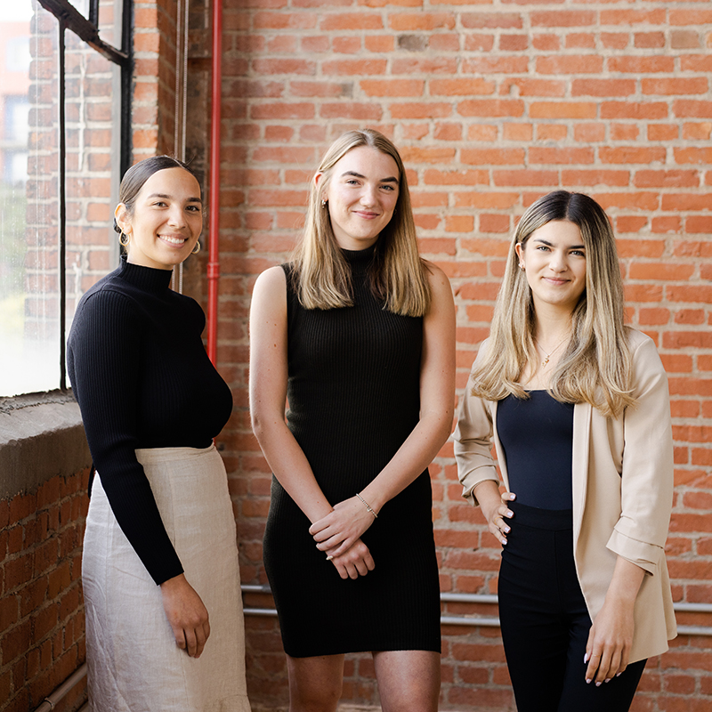 Three people standing side by side in front of an exposed brick wall, dressed in professional attire.