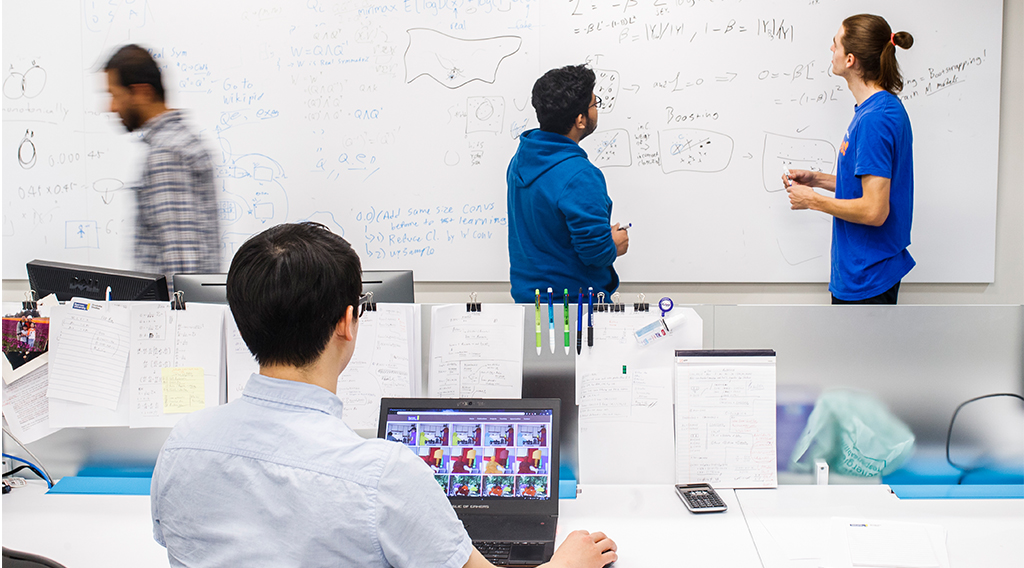 Students working on a white board while a student is working on a laptop in the foreground