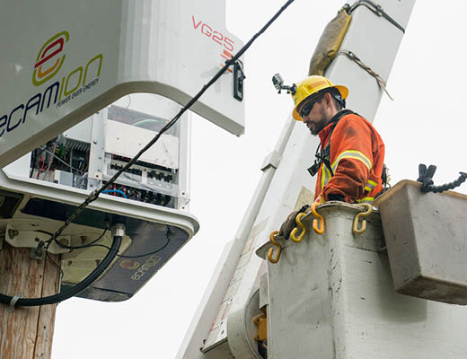 A male with an orange safety suit and hat is on a lift, opposite to a pole with an electrical panel open.