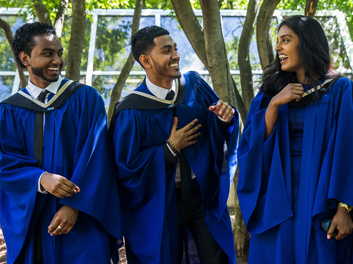 Three alumni, dressed in blue convocation robes, laugh together in the Quad.