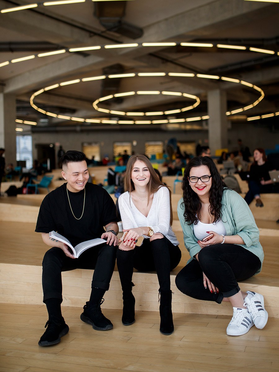 A group of three students smile at the camera while sitting under the ring lights in the SLC.