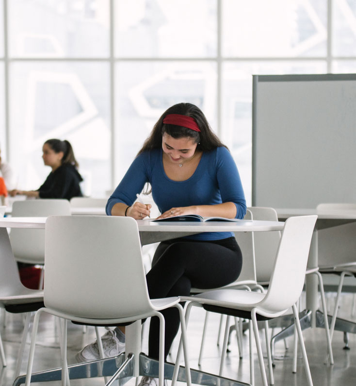 A female student sits alone at a table. She is writing on a piece of paper on the table while also holding open a book. Another student can be seen sitting at a different table in the background.