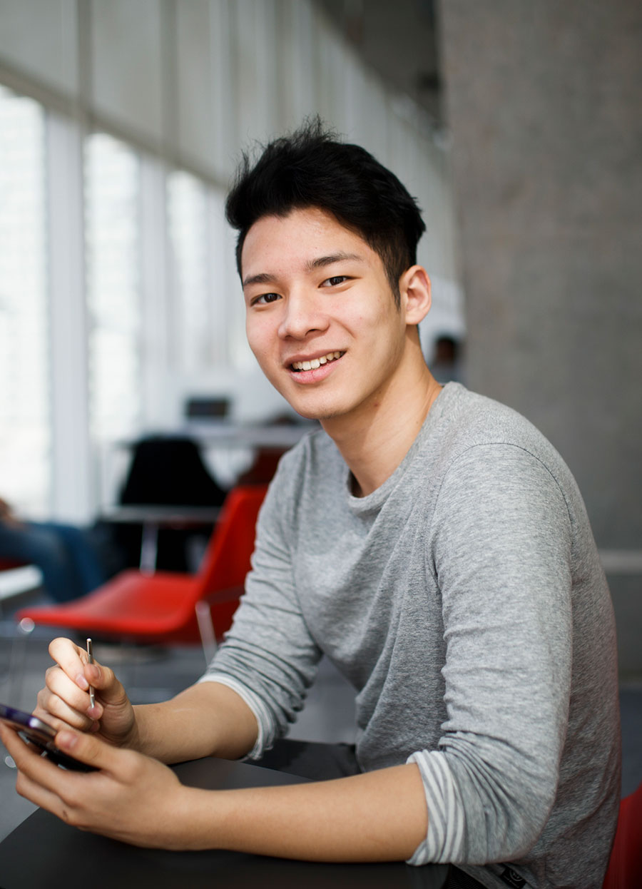 A student looking at the camera and smiling. He is holding a phone in his hand.