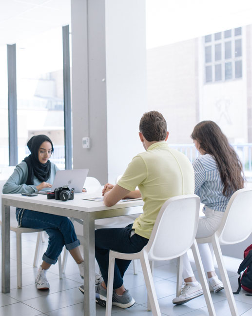 Three students sitting together at a table. Two students have their backs toward the camera. The third student is sitting across from them, typing on a laptop.