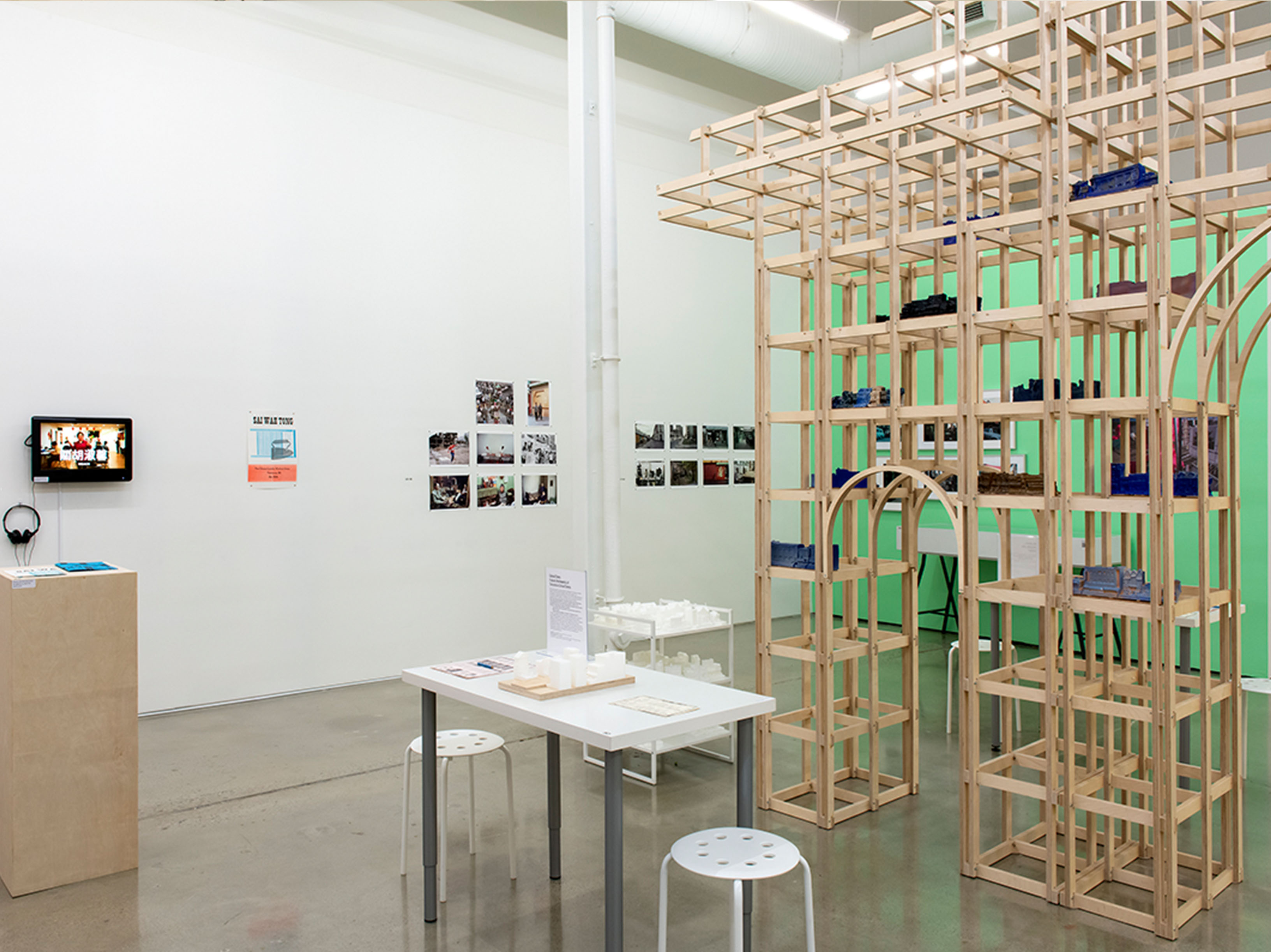 A gallery space with several objects on display. There is a wooden scaffold-looking structure, a tiered cart of small white objects, a white table with a sign and more white objects, and framed art on the walls.