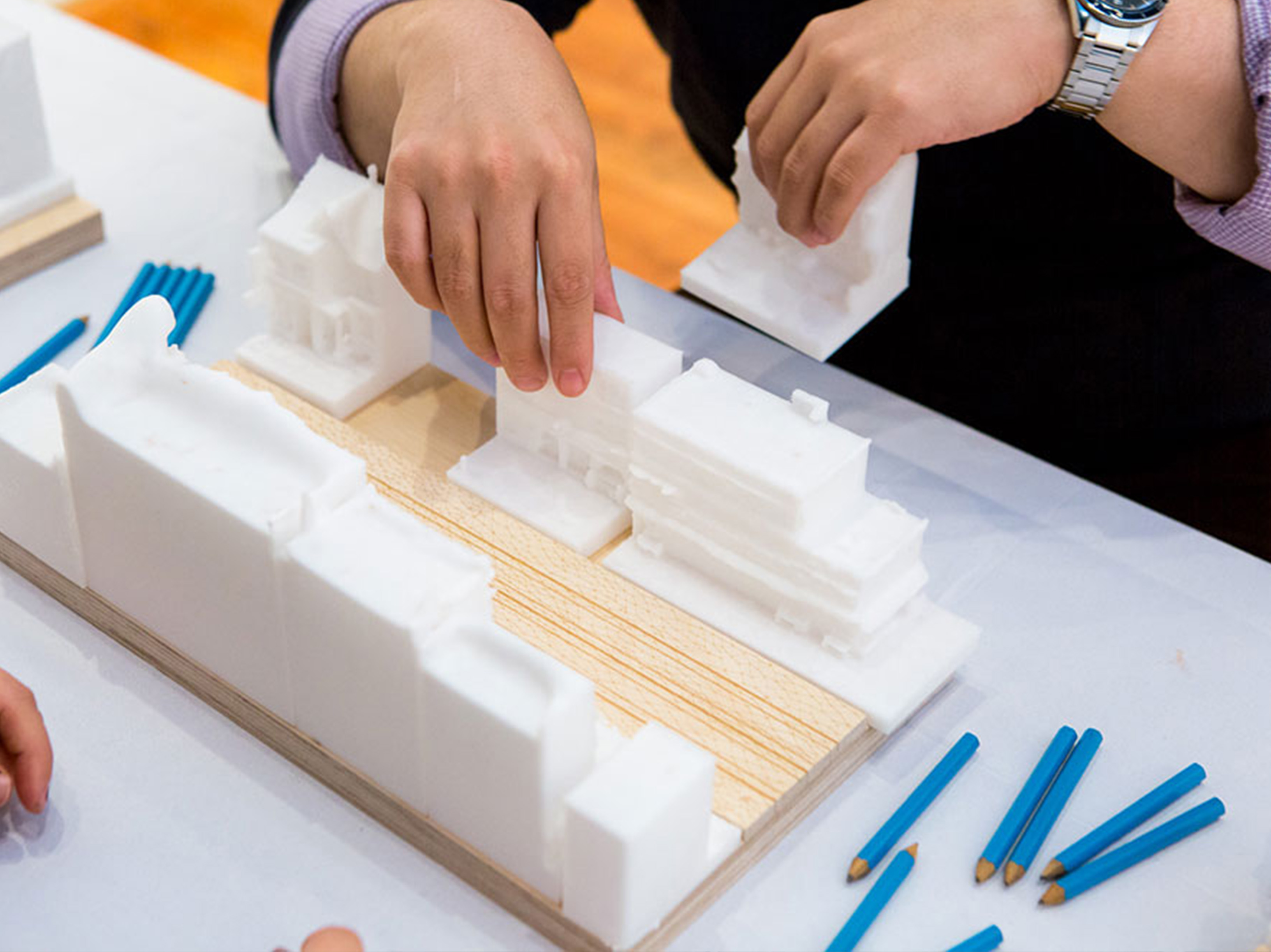 A close-up of hands assembling small white models of houses on a wood platform.