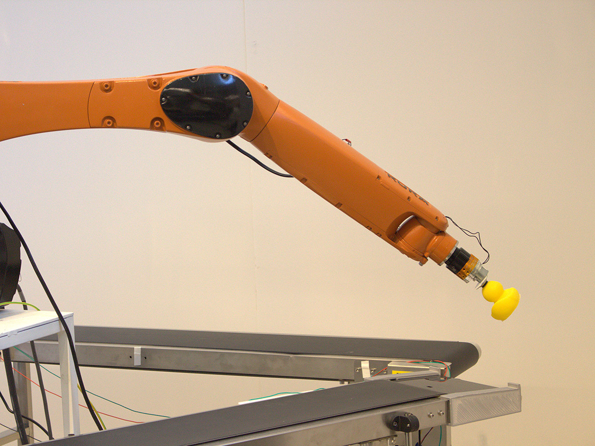 An orange robotic arm is extended and holding a yellow rubber duck over conveyor belts.