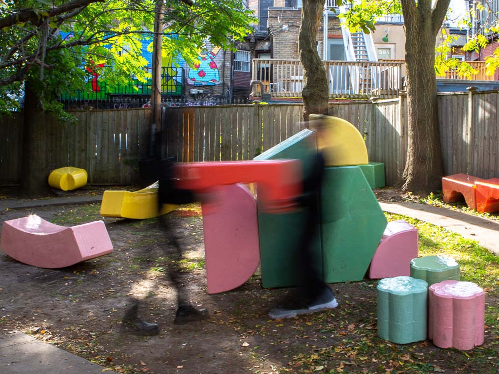 2 Figures have motion blur as they move throughout the brightly coloured shapes in a courta=yard. 