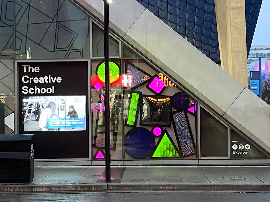A colourful window display of mirrors and lit geometric shapes beside a sign saying "The Creative School"