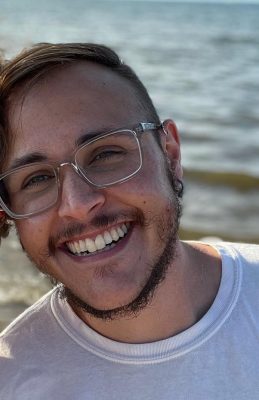 A digital photograph of a person with light skin, short dark hair styled with one side shaved, and a scruffy beard. They are wearing clear-framed glasses and a white t-shirt. They have a wide, joyful smile showing their teeth, and their eyes appear bright. The background features a body of water with gentle waves, slightly blurred. Sunlight illuminates their face, casting a warm glow. They are slightly angled towards the camera in a relaxed and happy pose.