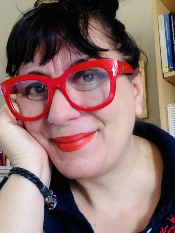 A digital photograph of a person with light skin, dark wavy hair styled in an updo, and bangs. They are wearing bold red-framed glasses, red lipstick, and a dark collared shirt. They have a warm, friendly expression with a slight smile and are resting their face on their hand. A black woven bracelet is visible on their wrist. The background features bookshelves filled with books and papers, suggesting an office or study setting. The lighting is soft and natural.