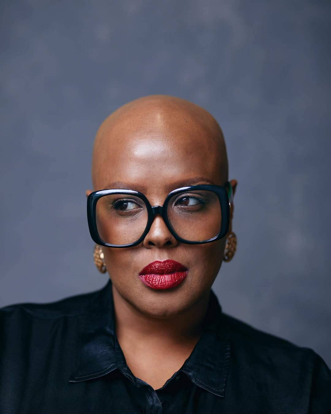 A digital photograph of a person with dark skin and a shaved head, wearing large black-framed glasses, gold earrings, and bold red lipstick. They have a confident expression and are looking slightly to the side. They are dressed in a black collared top. The background is a solid, muted gray color, and the lighting is soft and even.