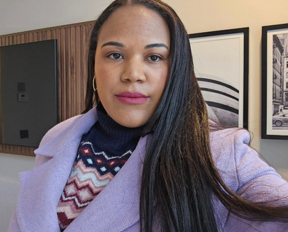A digital photograph of a person with dark skin and long, straight dark brown hair, wearing a lilac-colored coat over a patterned turtleneck sweater with zigzag designs in shades of navy, red, and white. They have a calm and confident expression, with subtle makeup including soft pink lipstick. They are wearing gold hoop earrings. The background features a modern interior with wooden paneling and framed black-and-white photographs of cityscapes on the wall. The lighting is soft and natural.