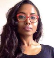 A digital photograph of a person with warm toned dark skin and long, wavy dark hair wearing red-framed glasses and a black top. They have a confident expression with a slight tilt of the head and are looking directly at the camera. They are wearing minimal makeup. The background is plain and light-colored. The lighting is soft and even.