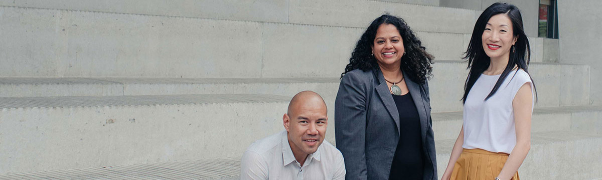 Three diverse TMU faculty members posing outside and smiling. Two are standing and one is sitting down on the steps.