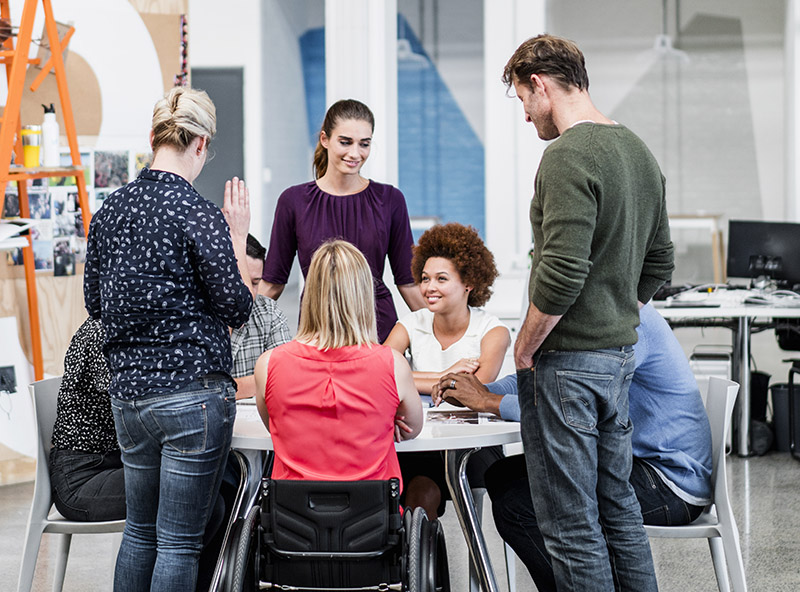 A group of eight diverse students gathered around a table to work on a project together. One student is using a wheelchair.