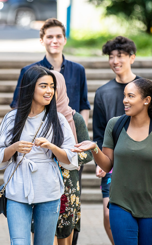 Five diverse students walking outside. Two students in the foreground are talking. 