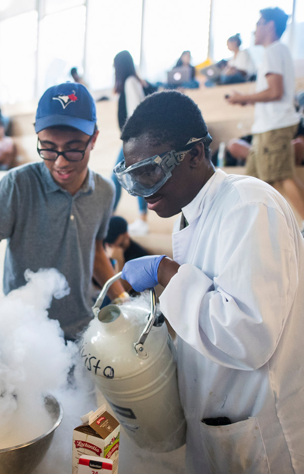 A number of diverse students in a large room. Two Black students in the foreground are doing a science project with smoke. 