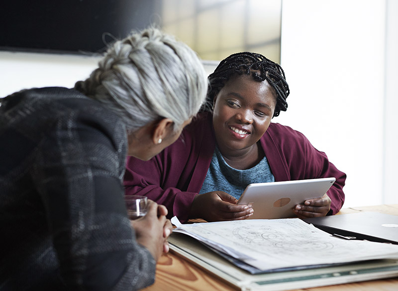 A Black student is seated at a table with a tablet device, while speaking with a racialized person with braided grey hair.