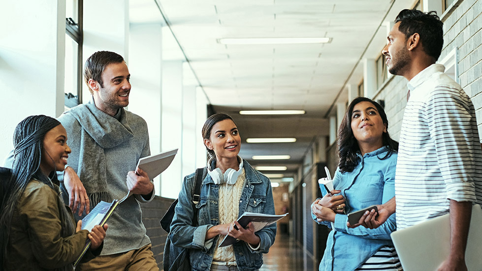 Five diverse students, standing and talking in the hallway of a building, holding notebooks and laptops.