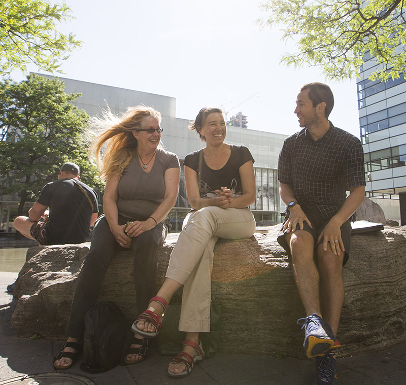 Three Indigenous students smiling and talking together by Lake Devo, with the Ryerson Image Centre and G. Raymond Chang School In the background.