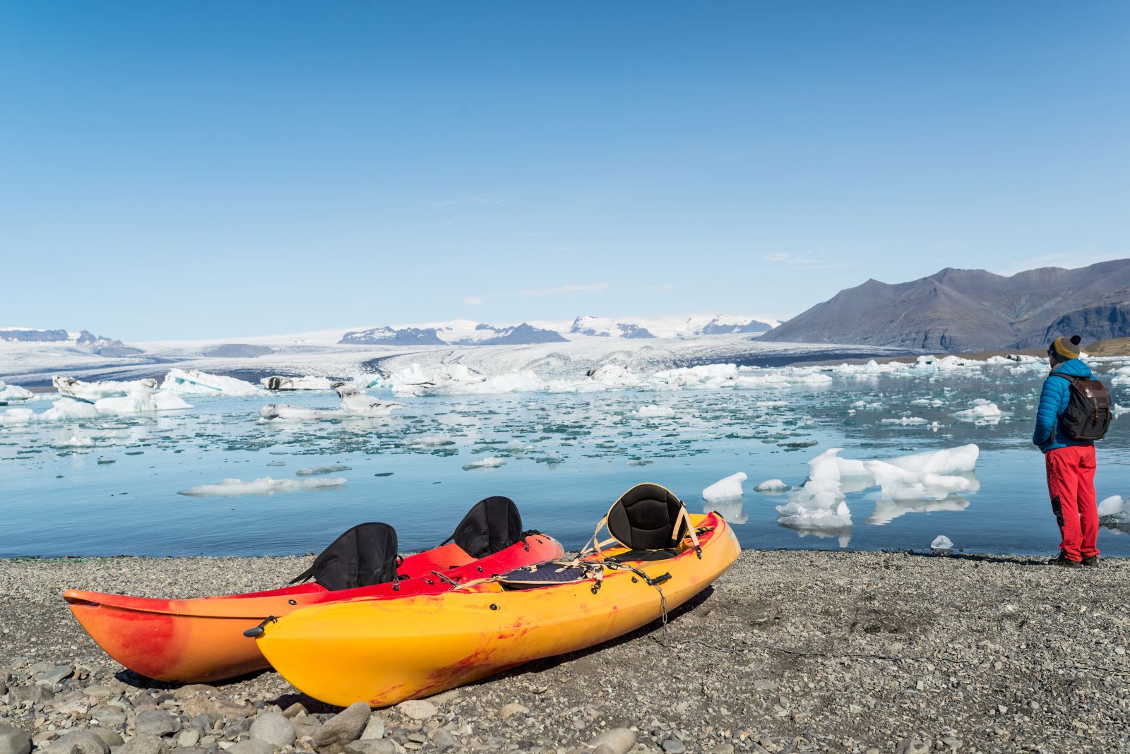 Three kayaks on the shore of a frozen icy lake, while one person stands by the shore.
