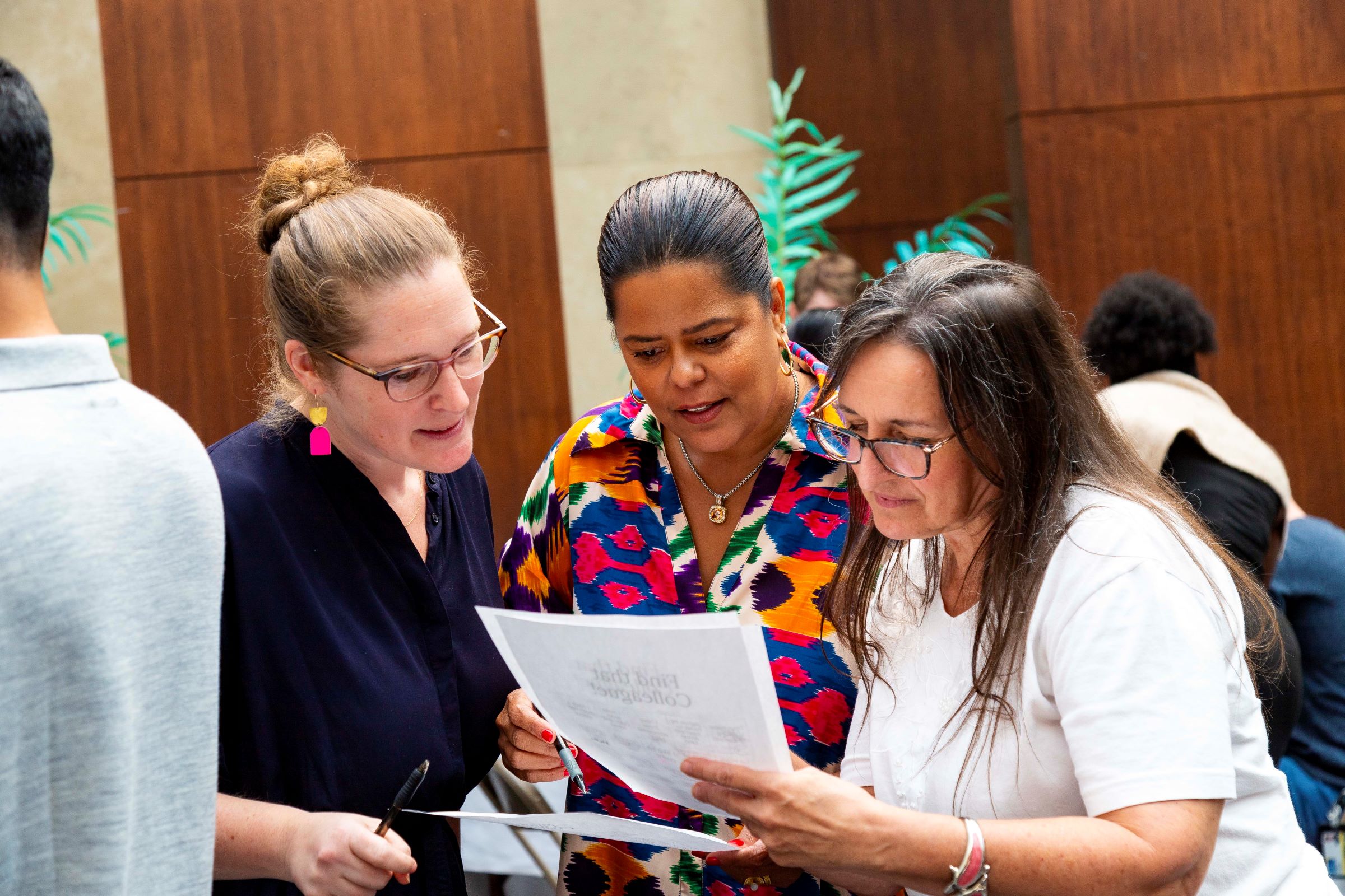 Three women are working together, huddled around a piece of paper as part of a trivia game. 