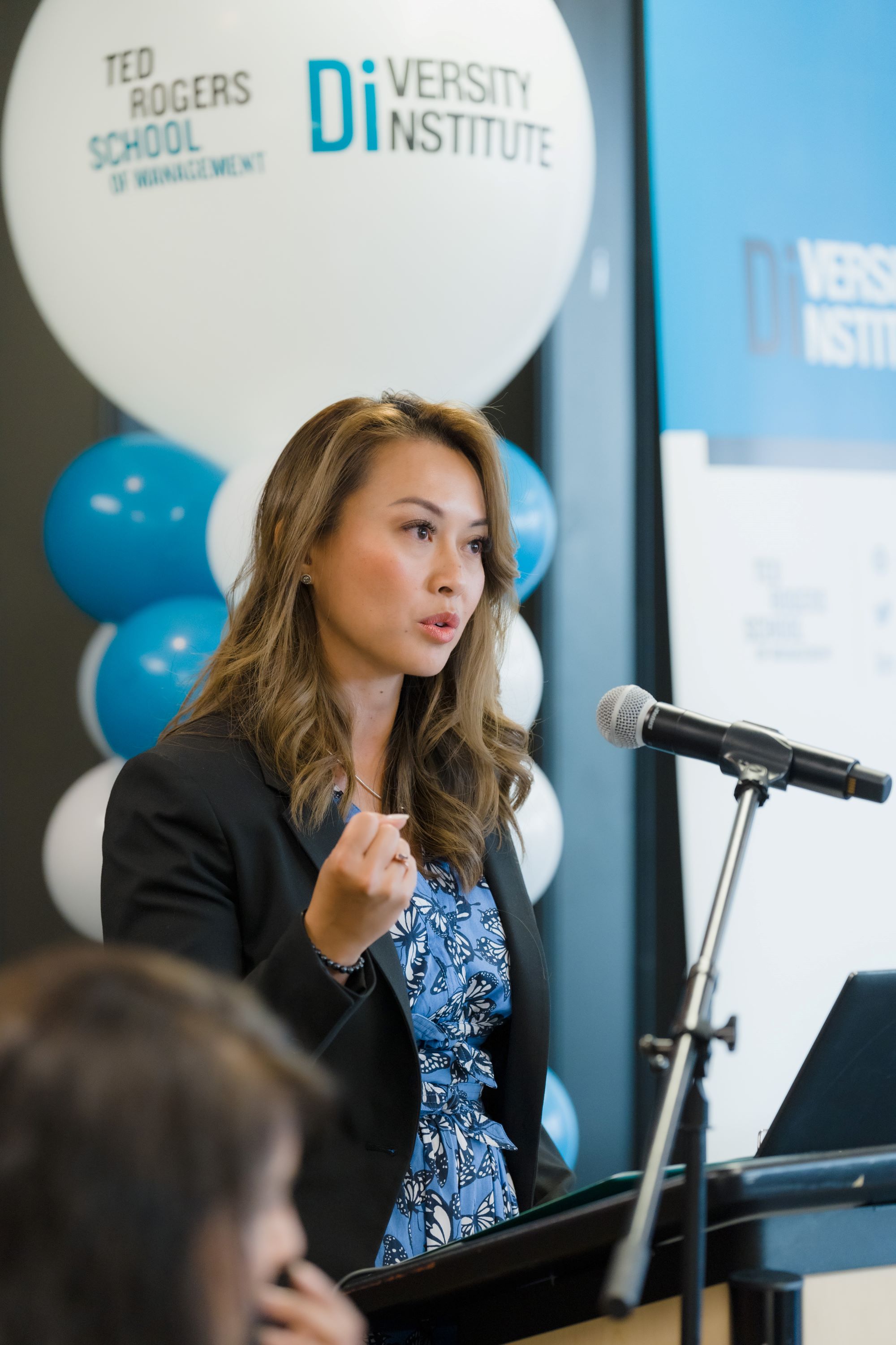 A Filipino woman wearing a blazer and blue dress, speaks into a microphone at a podium with blue and white balloons behind her.