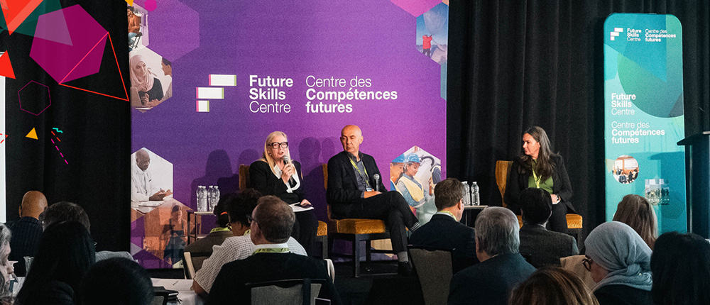 Three speakers address a large crowd at a bustling conference. 