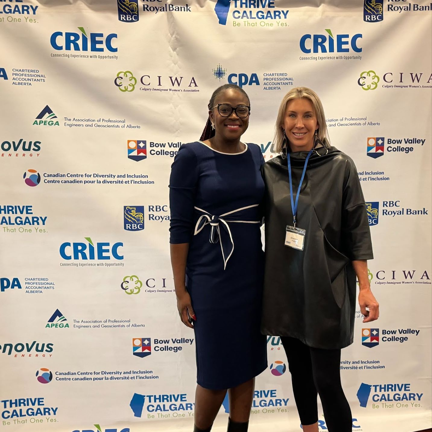 A Black woman and white woman smile together in front of a background showcasing a number of sponsors.