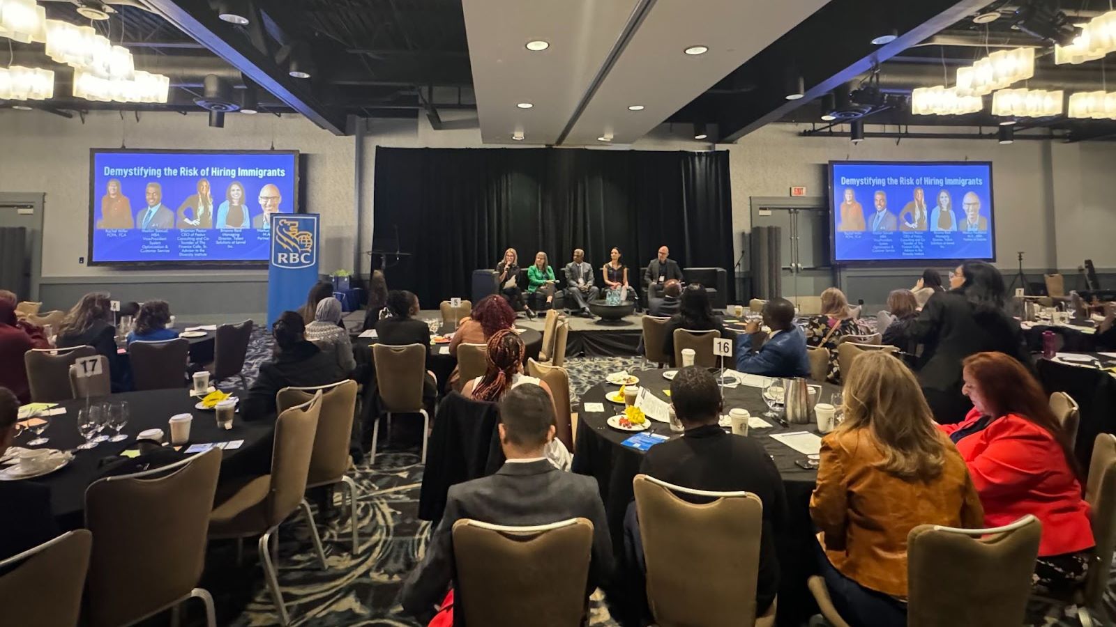 In a large conference room with many attendees, a pannel takes place on a stage with four speakers.