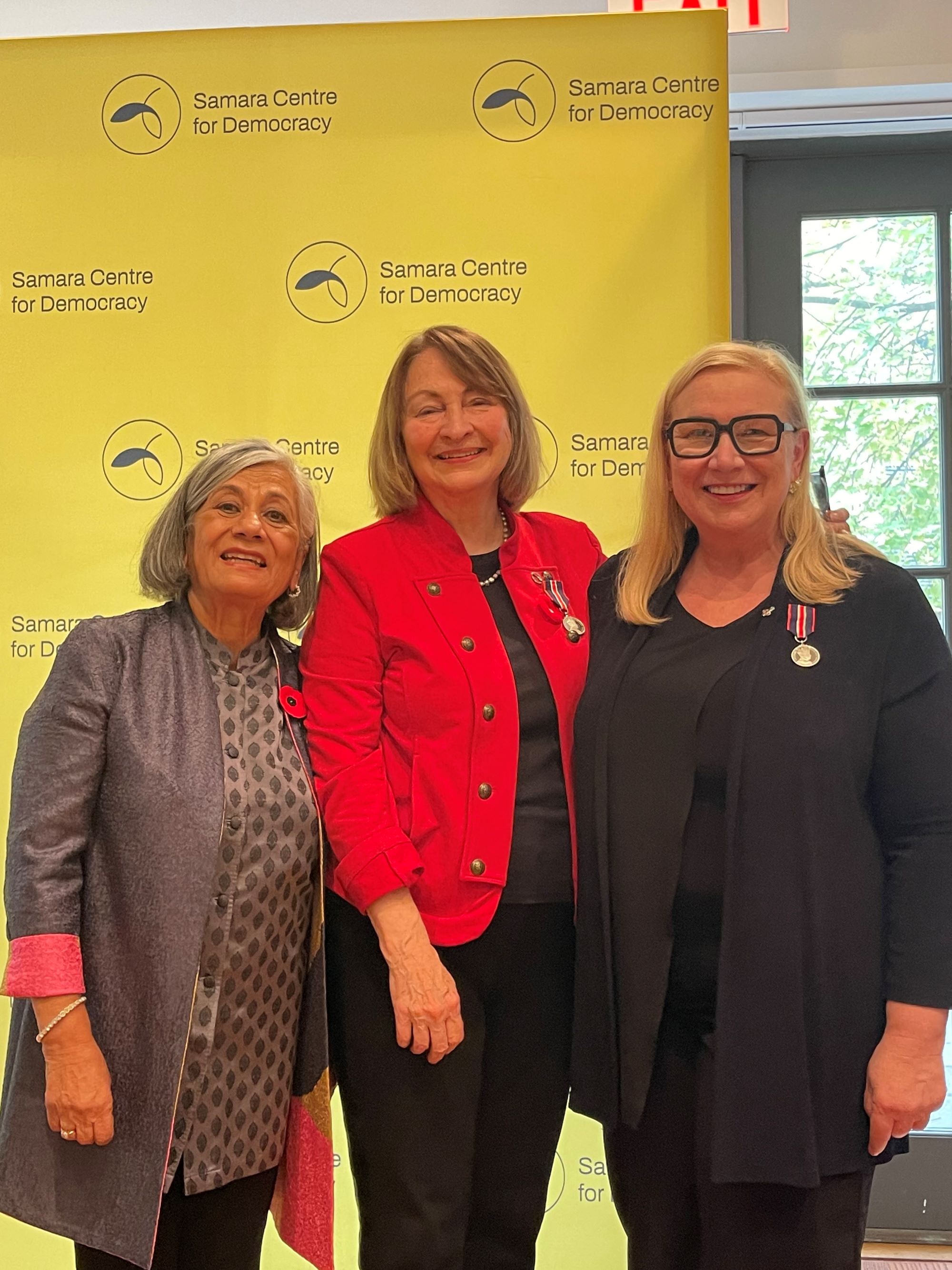 Three women standing and smiling for the camera in front of a backdrop that says Samara Centre for Democracy.