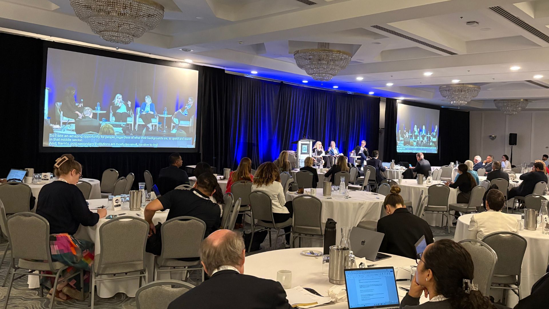 A group of people are seated in a large conference room, facing a stage with 4 people who are a part of a panel discussion. 