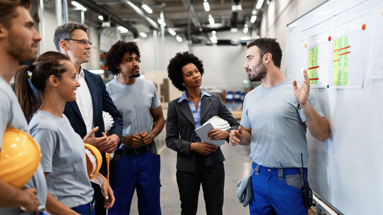 A diverse group of individuals speaking in front of a white board in a factory