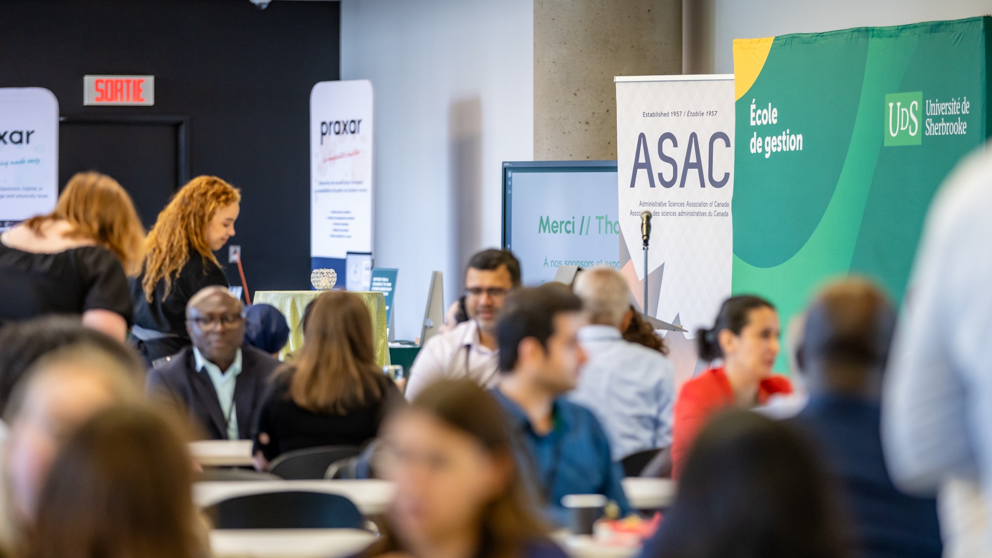 Dozens of people interact at a conference. Signs of different organizations can be seen in the background.