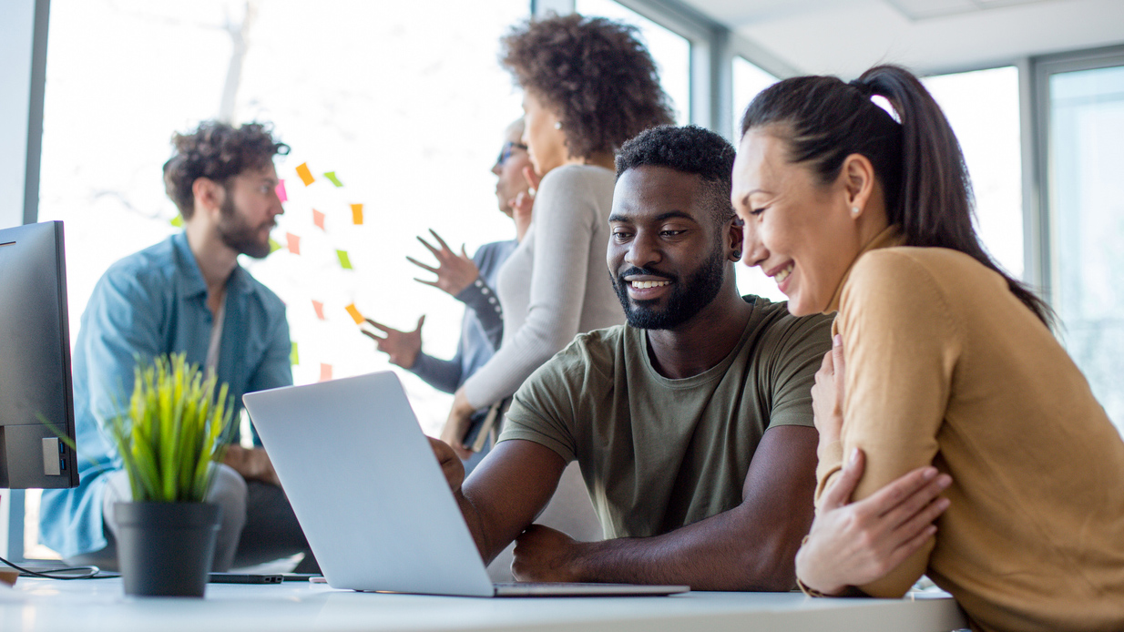 Two colleagues in an office looking at a laptop while another group meets in the background