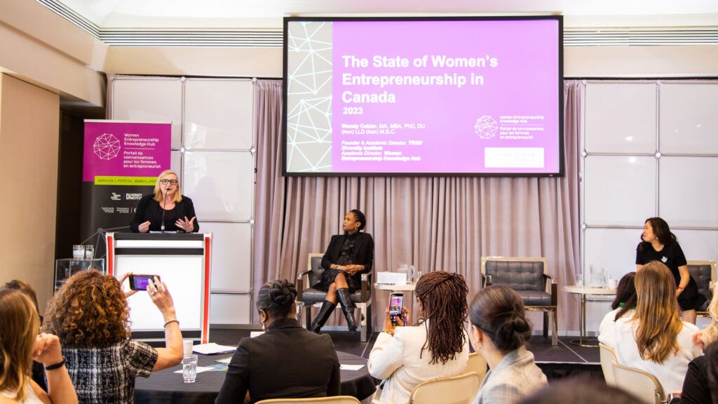 Three diverse women on stage where one woman is speaking behind the podium and the other two are sitting on chairs.
