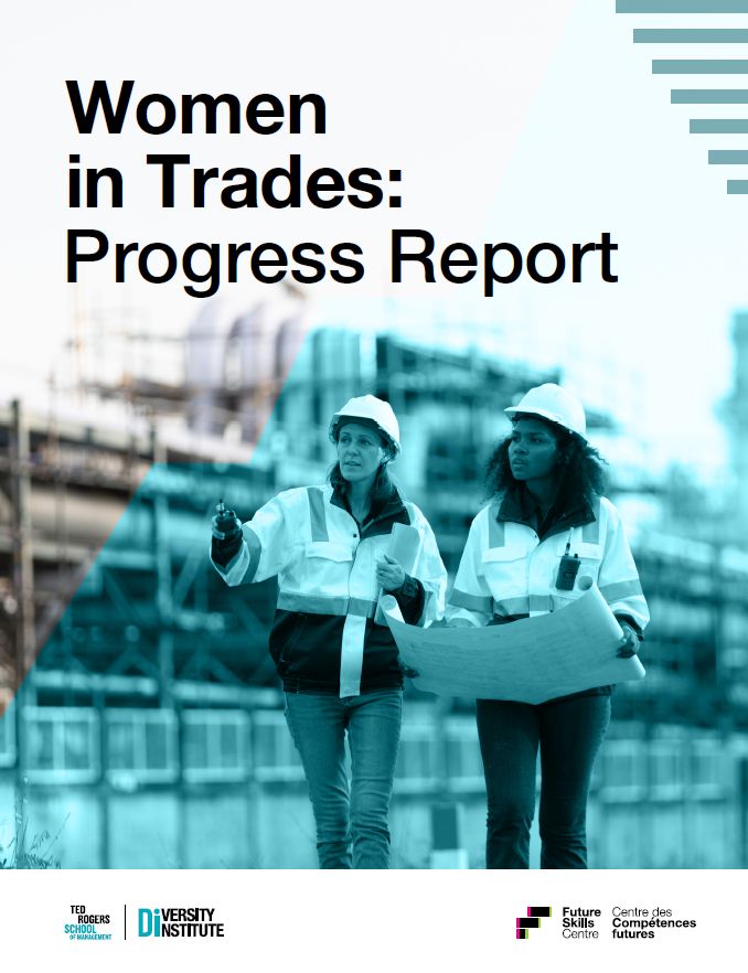 Two women in hard hats walking through a large industrial workplace while discussing blueprints.
