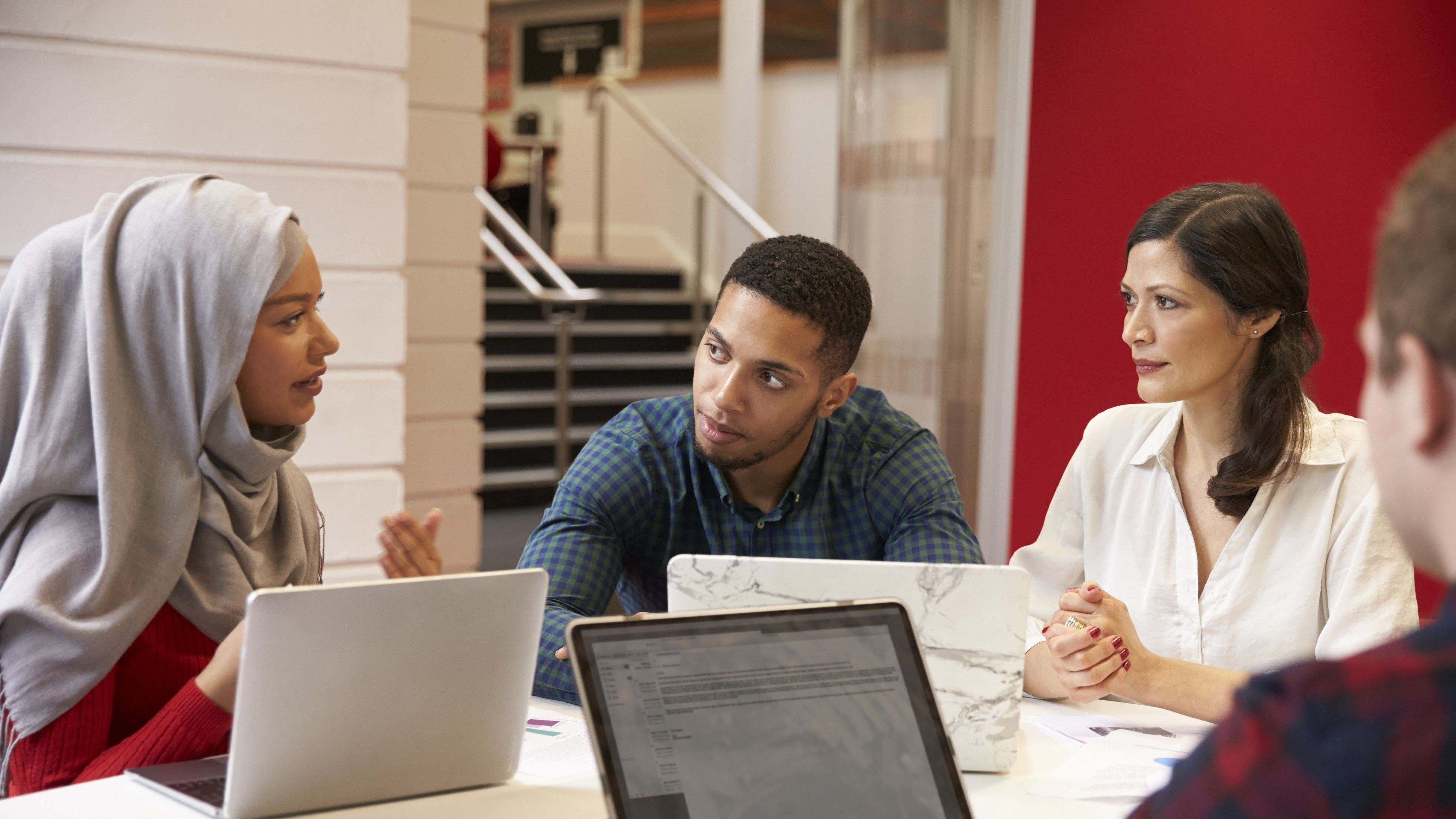 A group of diverse students meeting at a table with laptops and paperwork in front of them. 